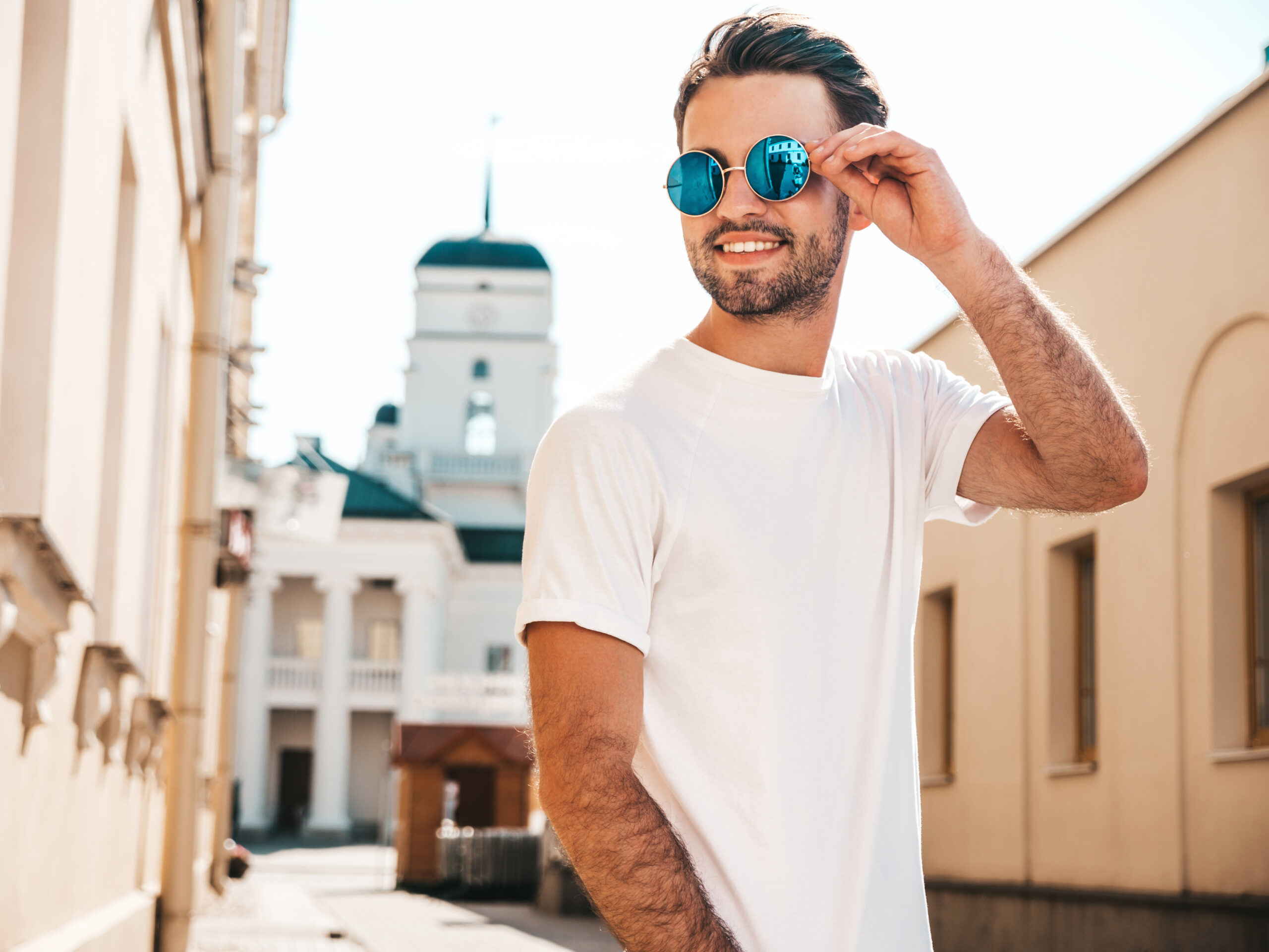 Portrait of handsome smiling stylish hipster lambersexual model.Man dressed in white T-shirt. Fashion male posing on the street background in round sunglasses